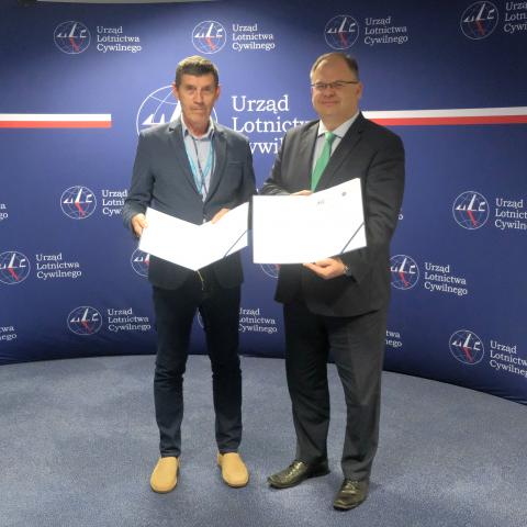 Prezes ULC i Przewodniczący PKBWL na tle ścianki z logo Urzędu Lotnictwa Cywilnego. W dłoniach trzymają podpisane porozumienie.